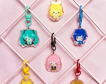 Könnte beinhalten: Sieben Schlüsselanhänger aus Acryl mit Cartoon-Charakter-Designs. Jeder Anhänger zeigt einen anderen Charakter mit unterschiedlichen Haarfarben, darunter Türkis, Gelb, Rosa und Braun. Die Anhänger sind an Metallverschlüssen in verschiedenen Farben befestigt, darunter Gelb, Blau und Rot.