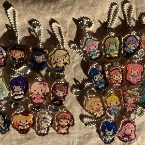 May include: Collection of acrylic keychains featuring colorful anime-style characters. Each keychain has a silver chain and a detailed illustration of a character with unique outfits and hairstyles. The keychains are arranged on a textured surface.