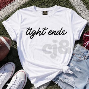 May include: A white short-sleeved t-shirt with a crew neckline and rolled sleeves. The front of the shirt features the text "tight ends" printed in a black cursive font. The shirt is made of a smooth fabric and has a knotted hem at the waist.