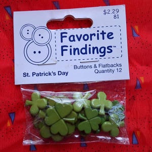 May include: A package of green shamrock-shaped buttons and flatbacks for St. Patrick's Day. The package is labeled "Favorite Findings" and contains 12 pieces. The background is a red fabric with blue and yellow triangle shapes.