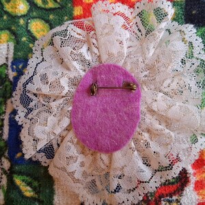 Puede incluir: Un broche de flor de encaje blanco con un centro de fieltro morado y un alfiler dorado en la parte posterior.