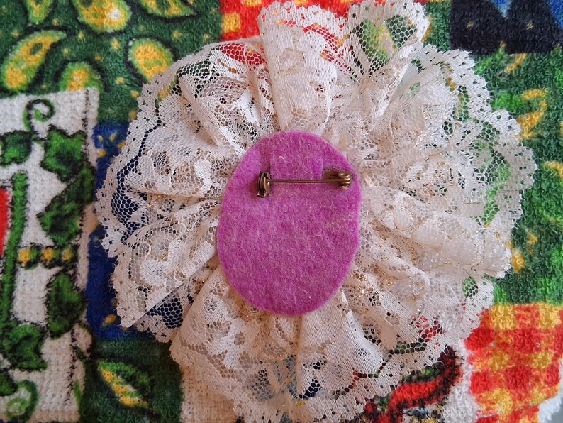 Puede incluir: Un broche de flor de encaje blanco con un centro de fieltro morado y un alfiler dorado en la parte posterior.