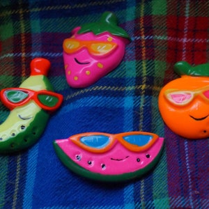 May include: Four colorful fruit-shaped figurines with smiling faces wearing sunglasses. The figurines are a pink strawberry, an orange orange, a green banana, and a pink watermelon.