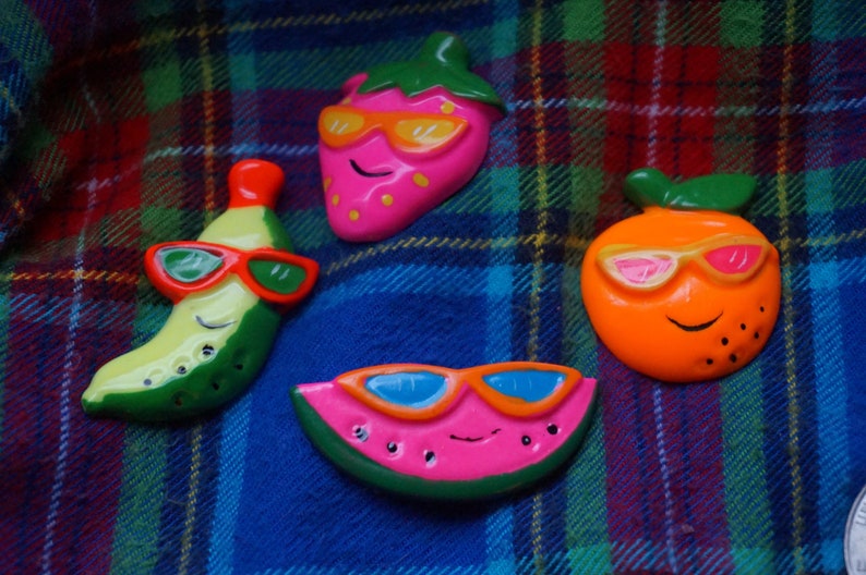 May include: Four colorful fruit-shaped figurines with smiling faces wearing sunglasses. The figurines are a pink strawberry, an orange orange, a green banana, and a pink watermelon.