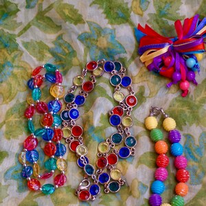 K&ouml;nnte beinhalten: Drei bunte Perlenketten auf einem floral gemusterten Stoffhintergrund. Eine Kette besteht aus gro&szlig;en, facettierten, mehrfarbigen Glasperlen. Eine weitere Kette besteht aus kleineren, runden, mehrfarbigen Glasperlen. Die dritte Kette besteht aus mehrfarbigen, runden Plastikperlen.