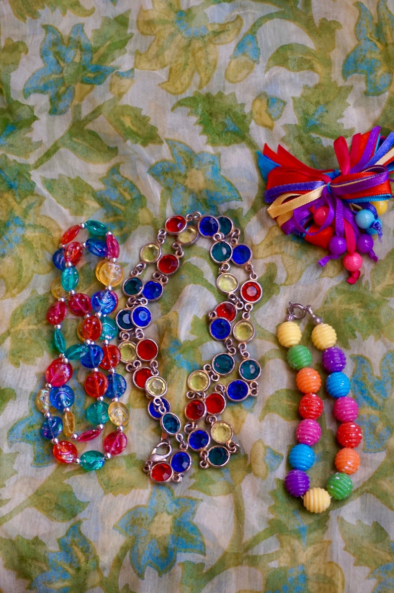 K&ouml;nnte beinhalten: Drei bunte Perlenketten auf einem floral gemusterten Stoffhintergrund. Eine Kette besteht aus gro&szlig;en, facettierten, mehrfarbigen Glasperlen. Eine weitere Kette besteht aus kleineren, runden, mehrfarbigen Glasperlen. Die dritte Kette besteht aus mehrfarbigen, runden Plastikperlen.