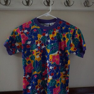 May include: A short-sleeved, purple t-shirt with a colorful floral print. The shirt has a crew neck and a ribbed collar. The print features pink, yellow, blue, green, and white flowers on a purple background. The shirt has the word "Ditango" printed on the front.