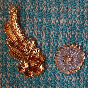 May include: Two gold and white decorative embellishments on a blue textured fabric. One embellishment is a gold leaf design with sequins. The other is a round white embellishment with gold sequins and a clear rhinestone in the center.