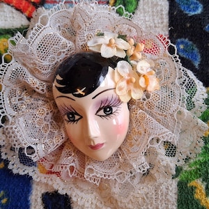 Puede incluir: Una cara de mu&ntilde;eca de porcelana con cabello negro y rasgos pintados, que lleva un cuello de encaje blanco con un adorno floral.