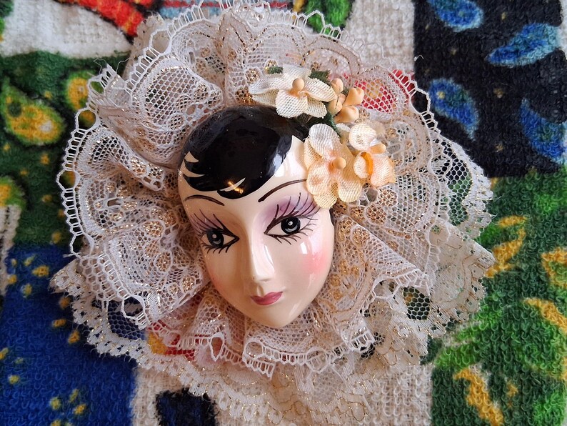 Puede incluir: Una cara de mu&ntilde;eca de porcelana con cabello negro y rasgos pintados, que lleva un cuello de encaje blanco con un adorno floral.