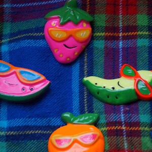 May include: Four colorful fruit-shaped pins with sunglasses. The pins are a watermelon, a strawberry, a banana, and an orange. The pins are on a plaid fabric background.