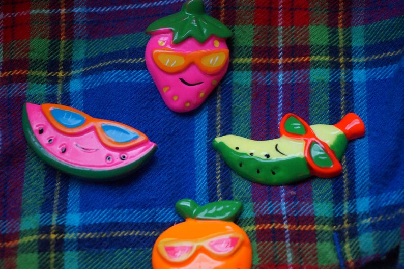 May include: Four colorful fruit-shaped pins with sunglasses. The pins are a watermelon, a strawberry, a banana, and an orange. The pins are on a plaid fabric background.