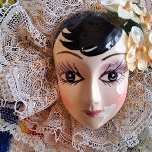 Puede incluir: Una cara de mu&ntilde;eca de porcelana con cabello negro, ojos pintados y un delicado cuello de encaje. La cara de la mu&ntilde;eca est&aacute; pintada con rubor rosa y tiene una suave sonrisa.