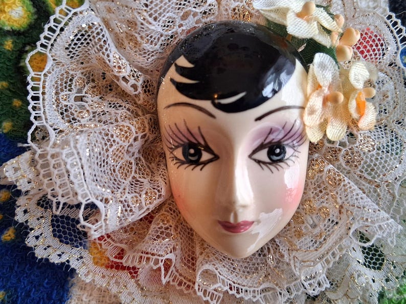 Puede incluir: Una cara de mu&ntilde;eca de porcelana con cabello negro, ojos pintados y un delicado cuello de encaje. La cara de la mu&ntilde;eca est&aacute; pintada con rubor rosa y tiene una suave sonrisa.
