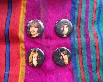 Könnte beinhalten: Vier runde Ansteckbuttons mit individuellen fotografischen Porträts der vier Mitglieder der Band The Beatles. Jeder Button hat einen schwarzen Hintergrund mit einem kontrastreichen Bild eines Musikers, angeordnet in einem zwei-mal-zwei Raster.