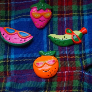 May include: Four colorful fruit figurines wearing sunglasses on a red and blue plaid background. The figurines are a watermelon, a banana, a strawberry, and an orange.