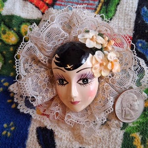 Puede incluir: Una cara de mu&ntilde;eca de porcelana con cabello negro y rasgos pintados, que lleva un cuello de encaje blanco con detalles florales.
