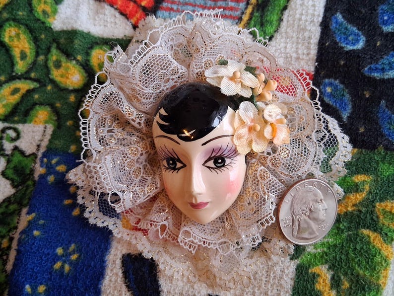 Puede incluir: Una cara de mu&ntilde;eca de porcelana con cabello negro y rasgos pintados, que lleva un cuello de encaje blanco con detalles florales.