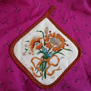 May include: A white cotton pot holder with a brown border and a floral design featuring a sunflower, poppy, and other flowers tied with a yellow ribbon. The text "C&B&D" is printed in the bottom right corner.