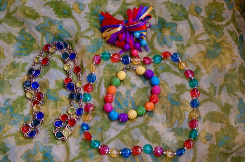 K&ouml;nnte beinhalten: Drei bunte Halsketten aus Glasperlen. Die Halsketten sind in einem Kreis auf einem floral gemusterten Stoff angeordnet. Die Halsketten bestehen aus verschiedenen Farben, darunter Rot, Blau, Gr&uuml;n, Gelb und Orange.