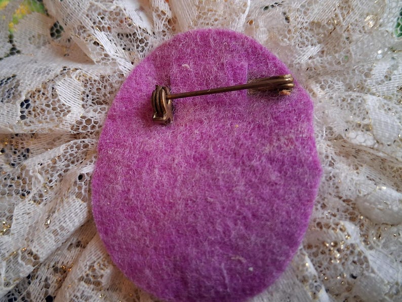 Puede incluir: La parte posterior de un broche de flor de fieltro morado con un alfiler de metal dorado.