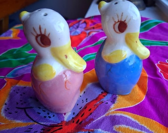 Puede incluir: Un par de saleros y pimenteros de cerámica con forma de pato. Un pato es rosa con detalles amarillos, el otro es azul con detalles amarillos. Ambos tienen cabezas blancas con ojos marrones y picos amarillos. Los saleros están sobre una superficie estampada colorida.