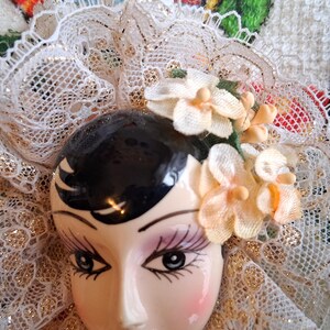 Puede incluir: Una cabeza de mu&ntilde;eca de porcelana con cabello negro y rasgos pintados, que lleva un velo de encaje blanco y una corona floral con flores rosas y blancas.