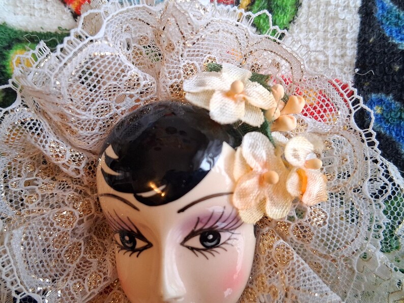 Puede incluir: Una cabeza de mu&ntilde;eca de porcelana con cabello negro y rasgos pintados, que lleva un velo de encaje blanco y una corona floral con flores rosas y blancas.