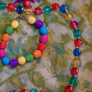 K&ouml;nnte beinhalten: Ein farbenfrohes Perlen-Halsketten- und Armband-Set mit abwechselnd gelben, gr&uuml;nen, blauen, roten und violetten Perlen. Die Halskette und das Armband liegen auf einem Stoff mit Blumenmuster.