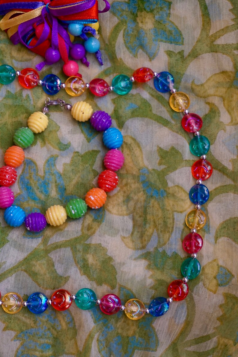 K&ouml;nnte beinhalten: Ein farbenfrohes Perlen-Halsketten- und Armband-Set mit abwechselnd gelben, gr&uuml;nen, blauen, roten und violetten Perlen. Die Halskette und das Armband liegen auf einem Stoff mit Blumenmuster.