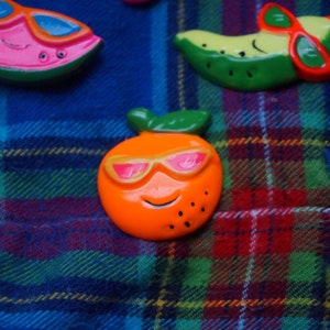 May include: Three colorful fruit-shaped magnets with sunglasses. A watermelon, a banana, and an orange.