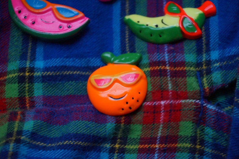 May include: Three colorful fruit-shaped magnets with sunglasses. A watermelon, a banana, and an orange.
