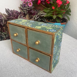 Könnte beinhalten: Kleine Holz-Aufbewahrungsbox mit vier Schubladen. Die Schubladen sind in einem gedämpften Grün mit goldenen Knöpfen lackiert. Die Seiten und die Oberseite der Box sind mit einem blau-grünen Blumenmuster verziert. Die Box steht auf einer grauen Oberfläche.