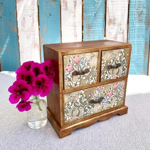 Könnte beinhalten: Kleine Holzschmuckschatulle mit drei Schubladen. Die Schubladen sind mit einem floralen Muster in Rosa, Blau und Grün verziert. Die Schatulle ist braun mit Schubladengriffen im antiken Stil. Links steht eine kleine Glasvase mit rosa Blumen.