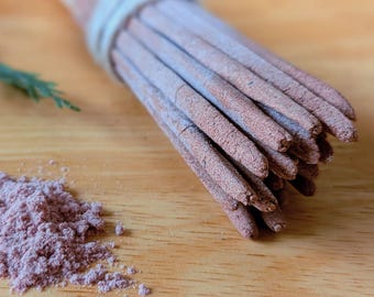 May include: A bundle of incense sticks tied with twine, with a pile of pink powder on a wooden surface. The incense sticks are a dusty rose color, and the powder is a similar shade. A sprig of green is in the background.