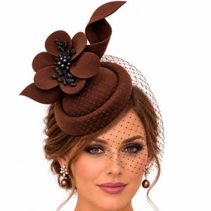 May include: A dark brown felt fascinator hat featuring a structured round base, a large matching felt flower with black beaded accents, and a delicate black netting veil that drapes over the front. The hat is worn with matching brown drop earrings.