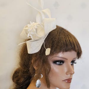 May include: A white feather and straw hat with a white floral accent. The hat is worn on a mannequin with brown hair.