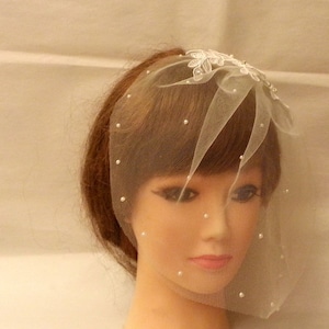 May include: A white bridal veil with a floral design and pearl accents. The veil is made of tulle and is designed to be worn over the face.