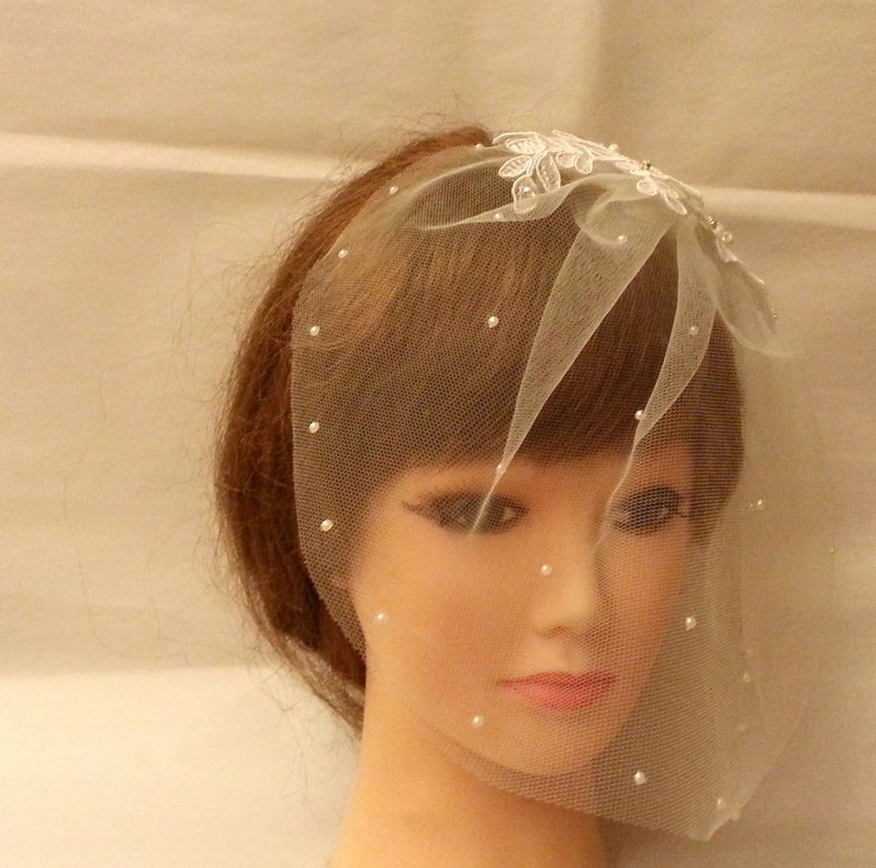 May include: A white bridal veil with a floral design and pearl accents. The veil is made of tulle and is designed to be worn over the face.