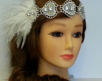 May include: A silver headband with a white feather accent. The headband is decorated with a row of silver hearts with rhinestones. The headband is worn on a mannequin with brown hair.