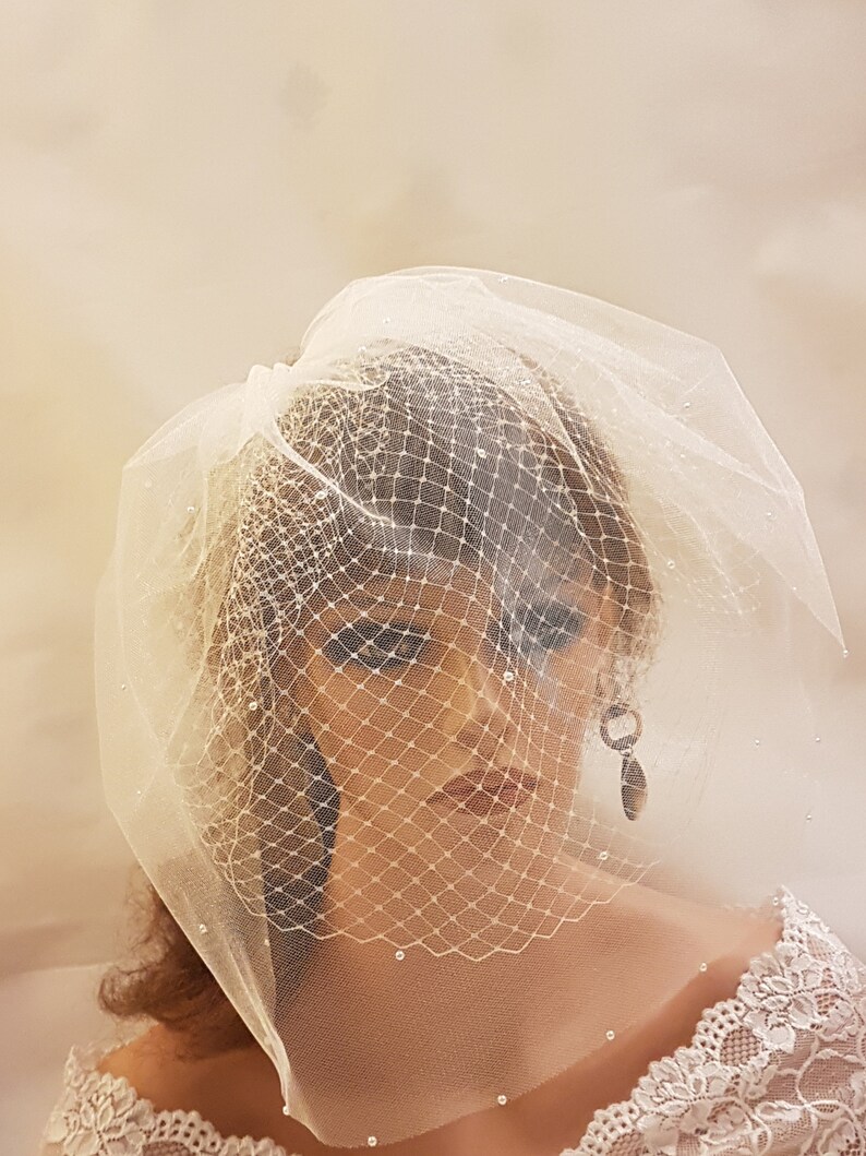 May include: A white bridal veil with a delicate net pattern and small pearl embellishments. The veil is gathered at the top and falls softly over the shoulders.