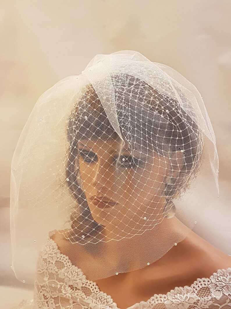 May include: A white bridal veil with a delicate net pattern and small pearl embellishments. The veil is short and covers the top of the head and part of the face.