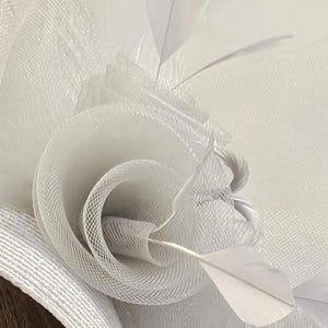 May include: White feather and tulle flower hat with a delicate, layered design. The hat is made of soft, white tulle and features a single white feather.