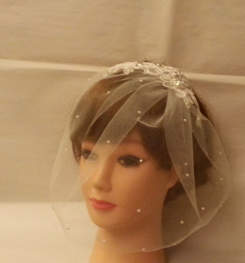 May include: A white bridal veil with a delicate lace trim and pearl embellishments. The veil is short and covers the top of the head.