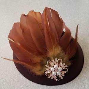 May include: A small brown felt hat with a brown and green feather trim and a gold and pearl brooch.