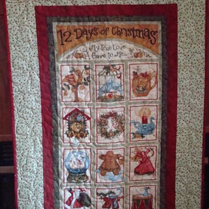 May include: A red, green, and brown quilted wall hanging with the text "12 Days of Christmas" and "My True Love Gave to Me..."  The hanging features 12 individual squares with illustrations of Christmas-themed items, such as a partridge in a pear tree, two turtle doves, and a French hen.