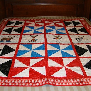 May include: A handmade quilt with a festive design. The quilt features alternating squares of black, white, and blue pinwheels, and red squares. The center squares have embroidered reindeer and snowflakes. The border is red with a white tree pattern.