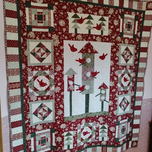 May include: A red, green, and white quilt with a birdhouse design. The quilt features a variety of patterns and textures, including floral prints, plaid, and stripes. The birdhouse is surrounded by birds and trees.