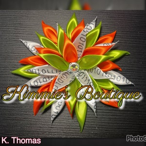 Peut inclure: Un accessoire pour cheveux en forme de fleur fait à la main, avec des rubans de satin orange, vert et blanc. Les rubans blancs portent des codes binaires imprimés. Un bijou étincelant est au centre. Le texte "Kimmie's Boutique" est en bas.
