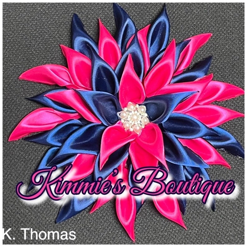 May include: A handmade, flower-shaped hair accessory. The petals are a combination of vibrant pink and navy blue satin ribbon, arranged around a pearl-centered embellishment. The text "Kimmie's Boutique" is visible.